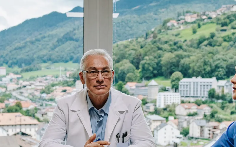 Erfahrener Arzt im weissen Kittel bei einer Besprechung, hinter ihm eine Schweizer Kleinstadt am Hang