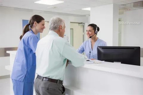 Medizinische Sekretärin am hellen Empfang eines Spitals in Zürich mit Headset, Pflegefachfrau bespricht kurz etwas, Bildschirm abgewandt