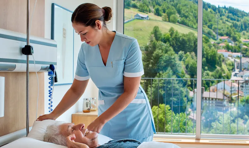 Eine Pflegefachfrau in hellblauer Berufskleidung richtet einem älteren Patienten im Spitalbett das Kissen.